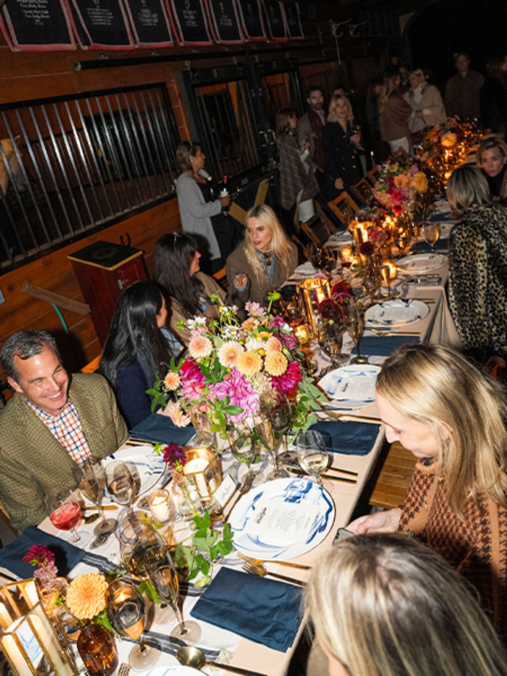 An elegant dinner party in a dimly lit restaurant with exposed brick walls and wooden beams. Guests are seated along both sides of a long table decorated with elaborate floral centerpieces featuring pink, orange, and yellow blooms. The table is set with white plates, navy blue napkins, wine glasses, and glowing candles creating warm ambient lighting. Approximately a dozen well-dressed attendees are engaged in conversation, with some standing and mingling in the background near orange-framed windows or panels.
