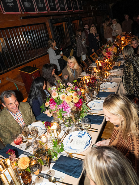 An elegant dinner party in a dimly lit restaurant with exposed brick walls and wooden beams. Guests are seated along both sides of a long table decorated with elaborate floral centerpieces featuring pink, orange, and yellow blooms. The table is set with white plates, navy blue napkins, wine glasses, and glowing candles creating warm ambient lighting. Approximately a dozen well-dressed attendees are engaged in conversation, with some standing and mingling in the background near orange-framed windows or panels.
