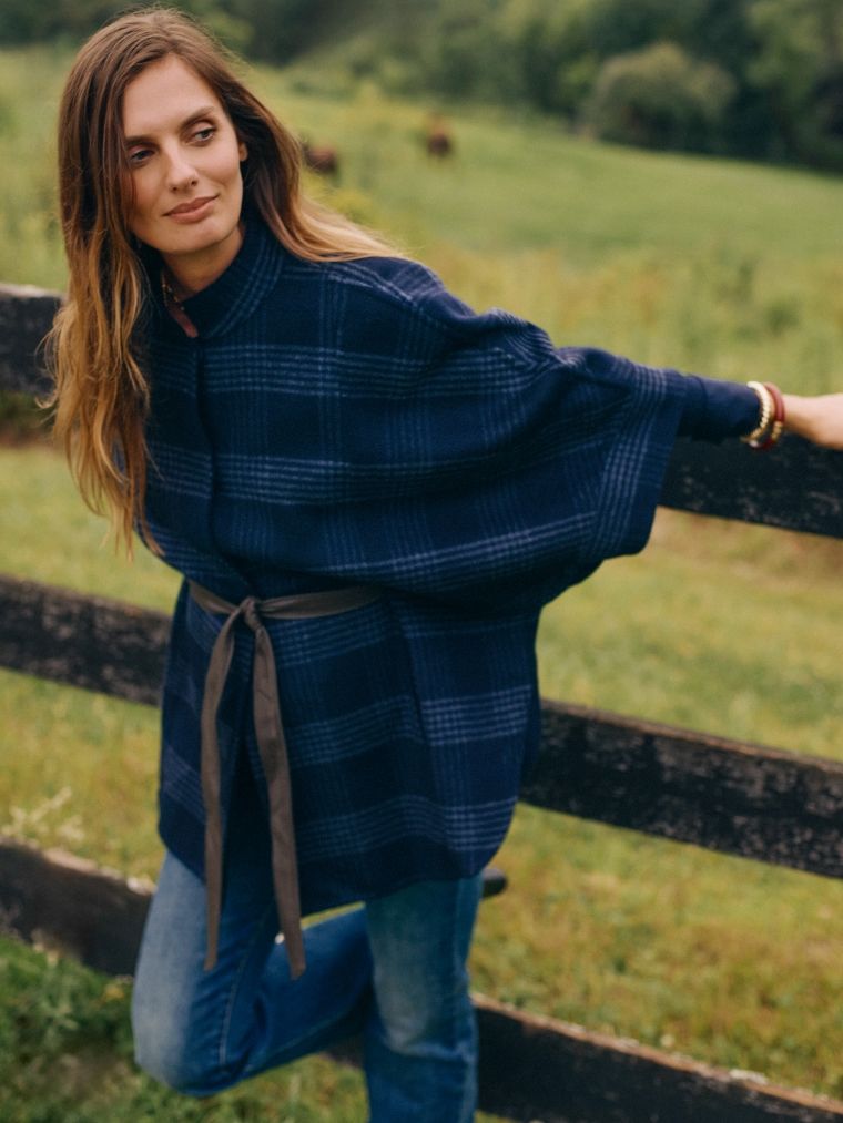 woman standing outside in field against a fence wearing a dark navy plaid cape