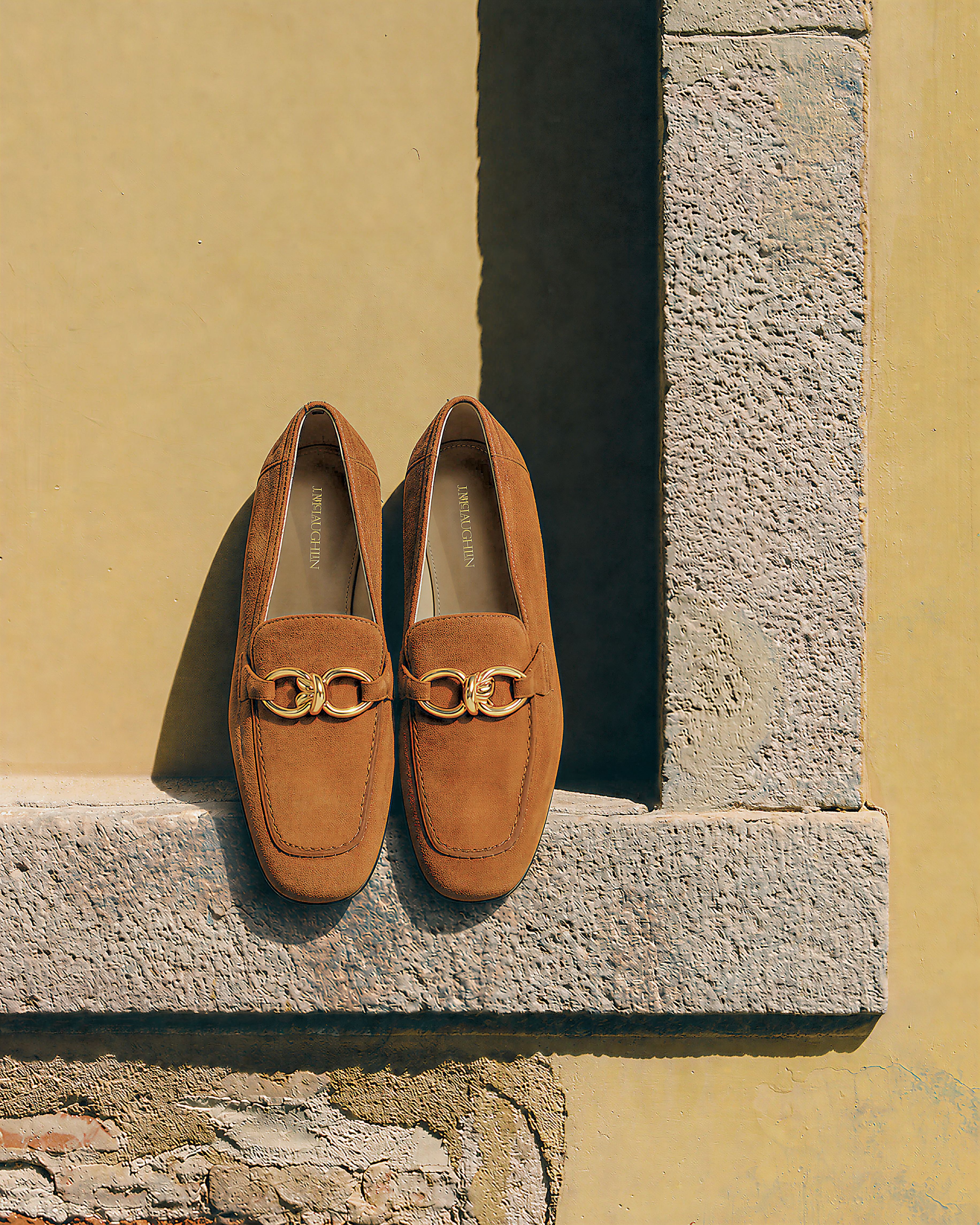 A pair of brown suede loafers with gold buckles sits on a stone ledge against a yellow wall.