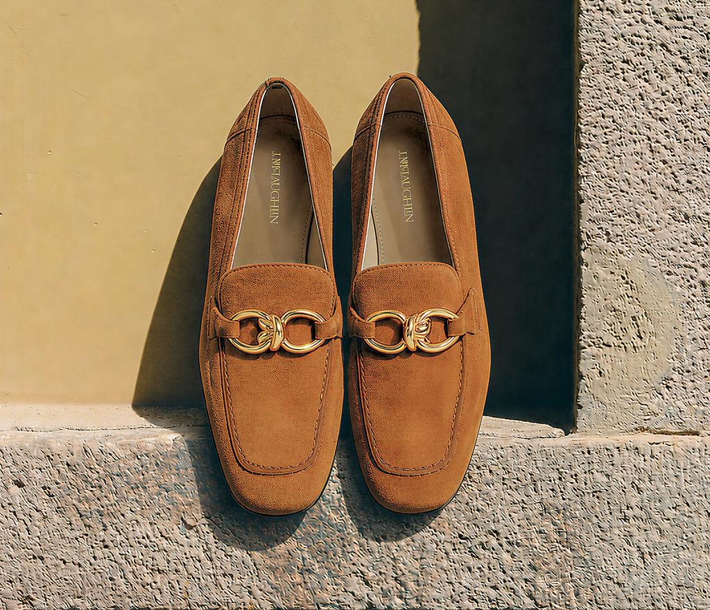 A pair of brown suede loafers with gold buckles sits on a stone ledge against a yellow wall.