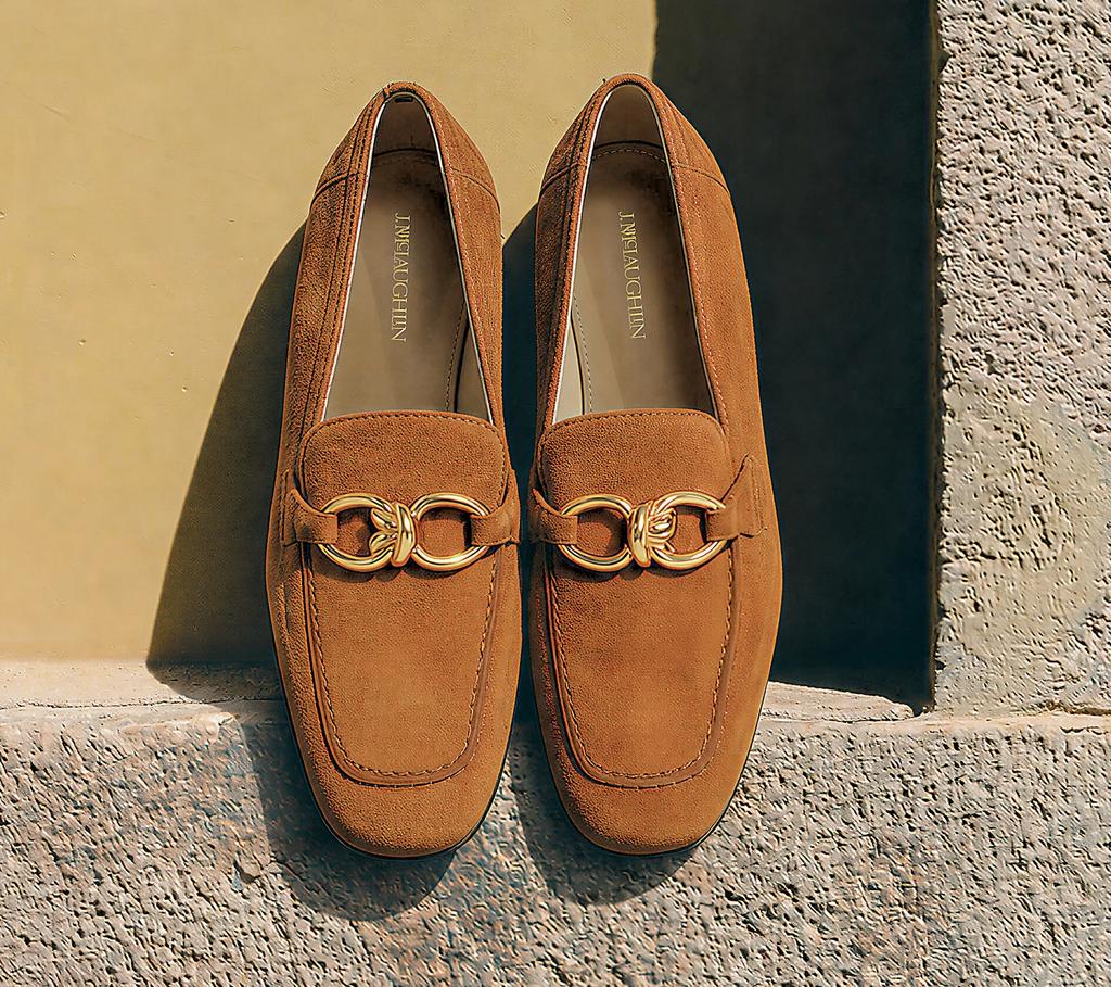 Brown suede loafers with gold buckles resting on a stone ledge against a yellow wall.