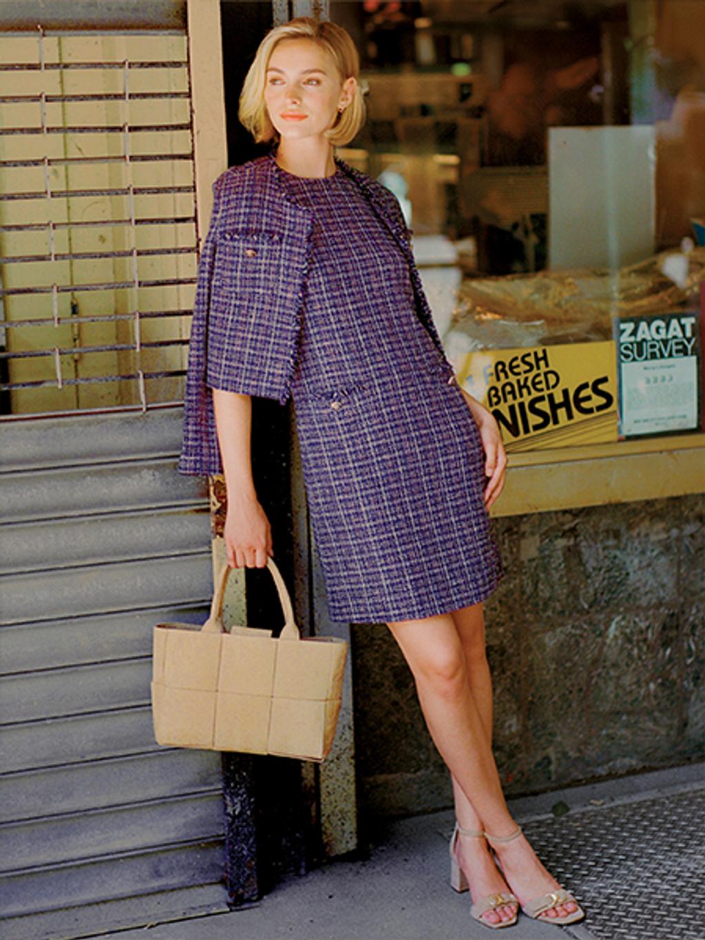 Women with short blonde hair leaning against a wall while wearing a purple tweed dress and jacket, holding a sand-colored handbag.