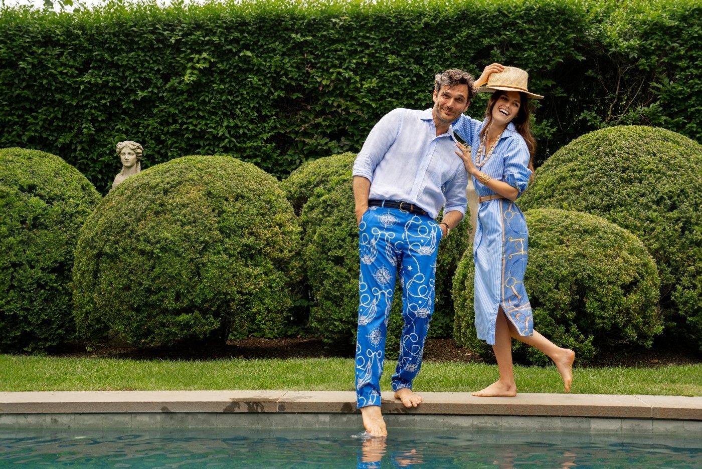 man and woman standing by a pong, she is wearing blue and white stripe shirtdress, he is wearing blue nautical print pants and blue linen shirt