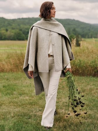 A woman in a plaid cape and cream pants walks through a field, holding a bundle of dark dried flowers.
