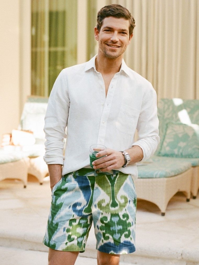 A smiling man in a white linen shirt and green and blue ikat-patterned shorts holds a drink outdoors.