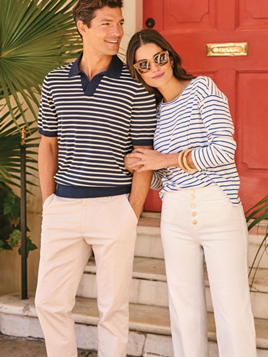 Male model in striped sweater and khakis, female model in striped tee and white jeans, both standing outside red door in Charleston
