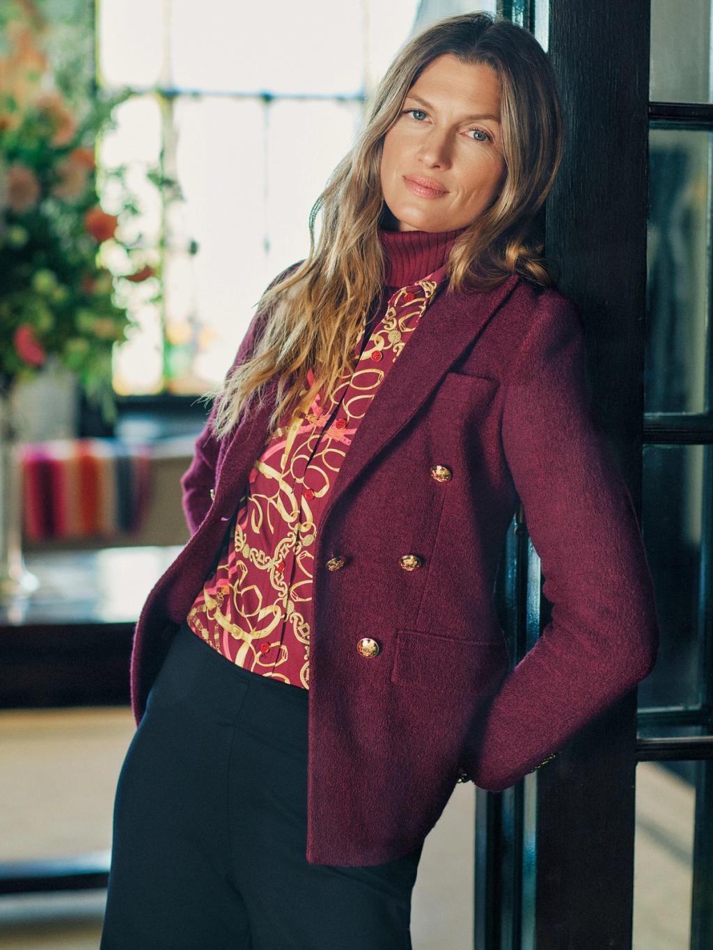Woman in a burgundy blazer, patterned shirt, red turtleneck, and dark pants leaning against a doorframe.