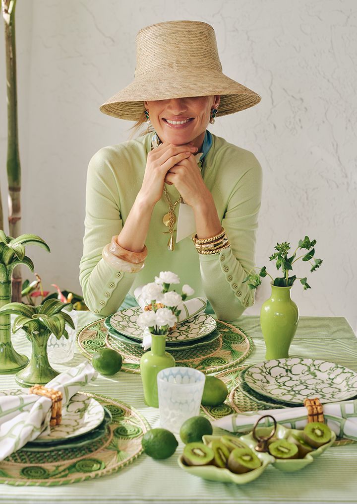 A smiling woman in a wide-brimmed straw hat sits at a vibrant green and white table, featuring patterned dishes, limes, sliced kiwis, and ceramic palm tree decorations.