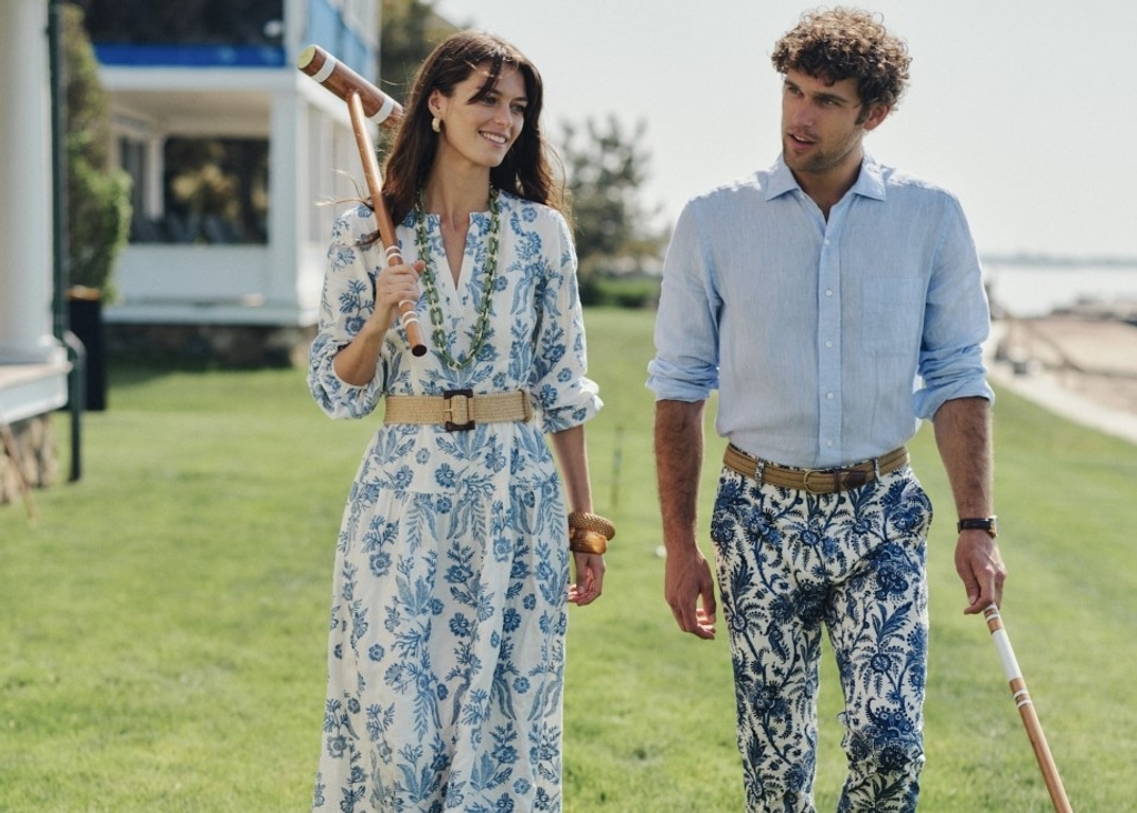 A woman in a blue and white floral dress and a man in a blue shirt and patterned pants walk on a lawn, each holding a croquet mallet.