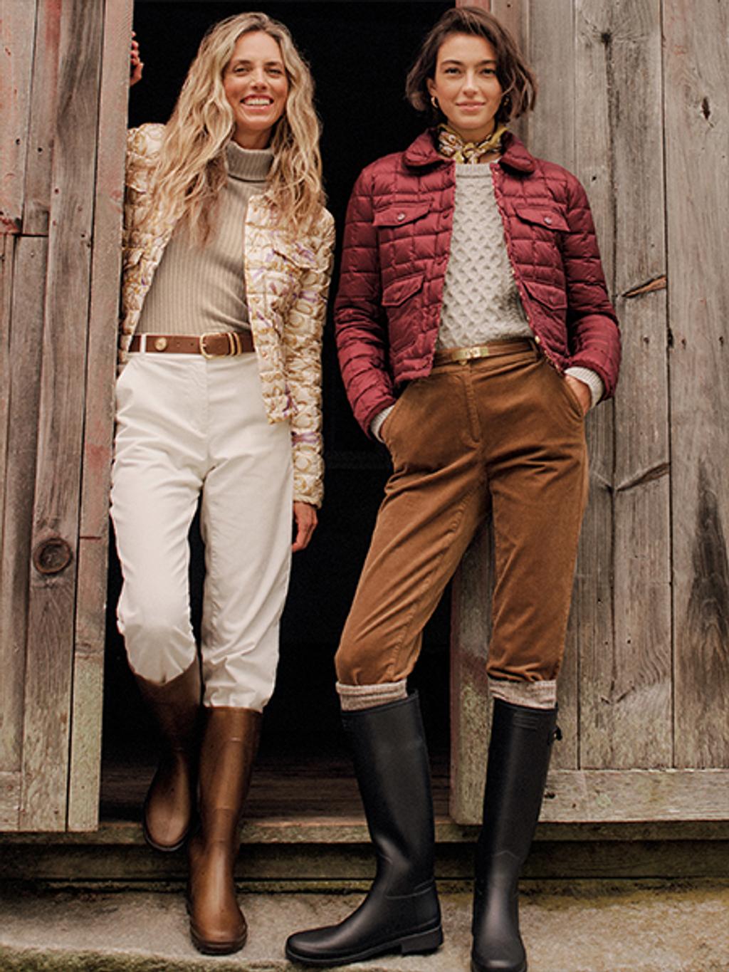 Two smiling women in quilted jackets, tailored pants, and boots stand in a rustic wooden doorway.