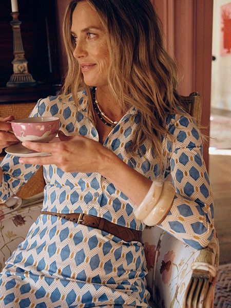 Female model in white/blue printed dress and brown belt posing with a coffee cup and saucer.