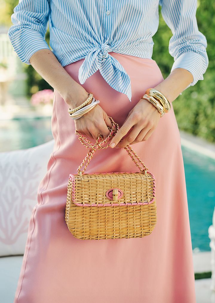 A person in a tied blue striped shirt and pink skirt holds a small straw bag with pink trim.