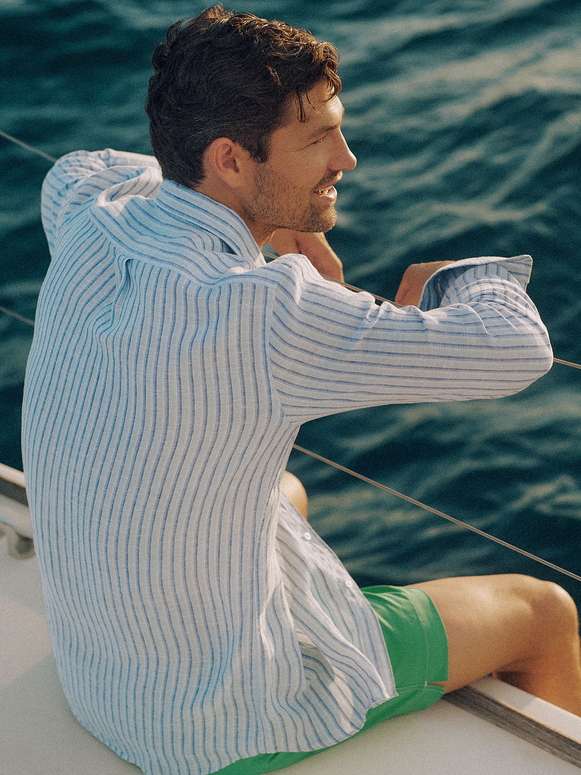 A smiling man in a striped shirt and green shorts looks out from a boat.