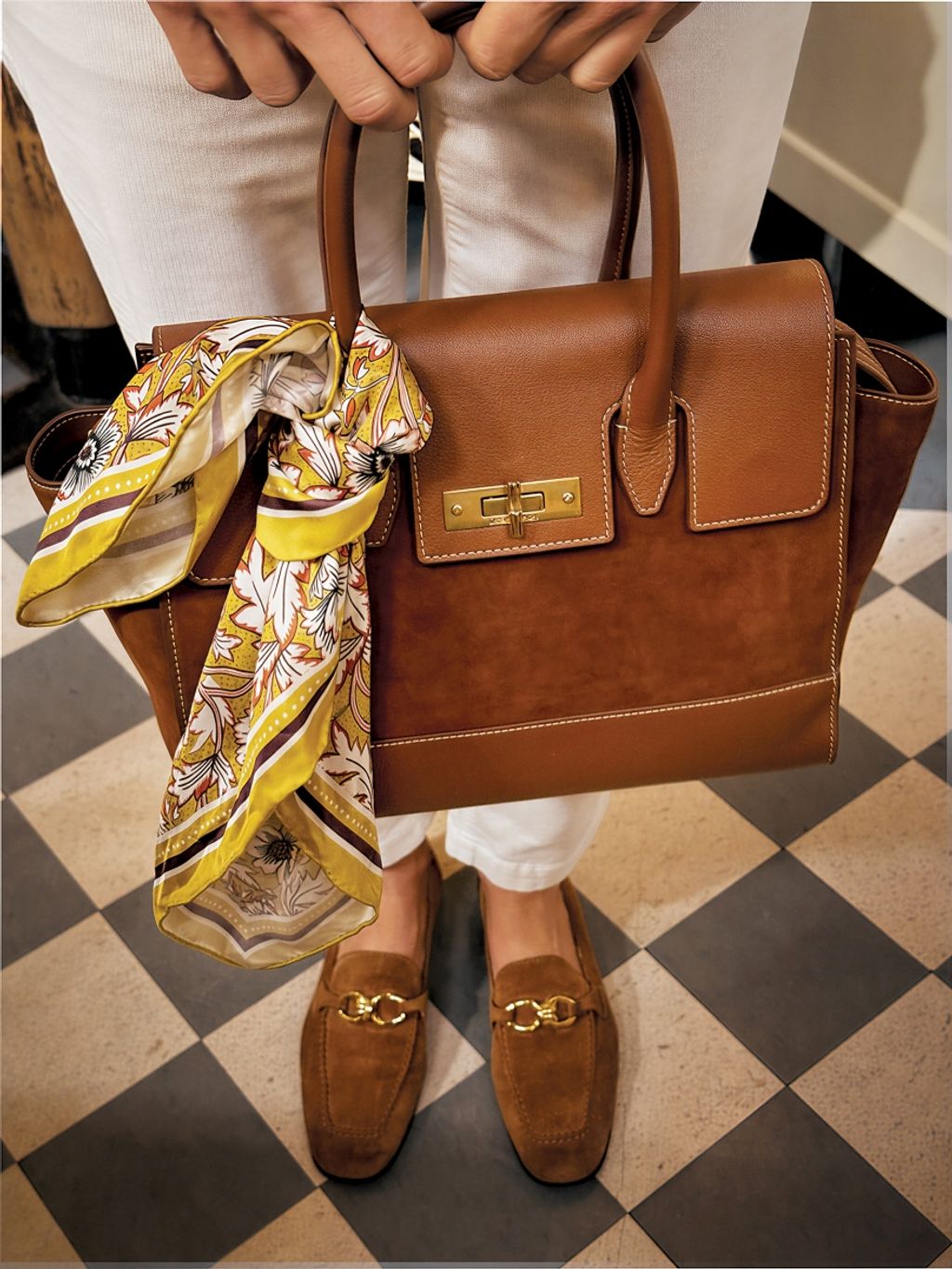 A person holding a brown leather and suede handbag with a yellow floral scarf, wearing white pants and brown suede loafers with gold buckles, on a checkered floor.