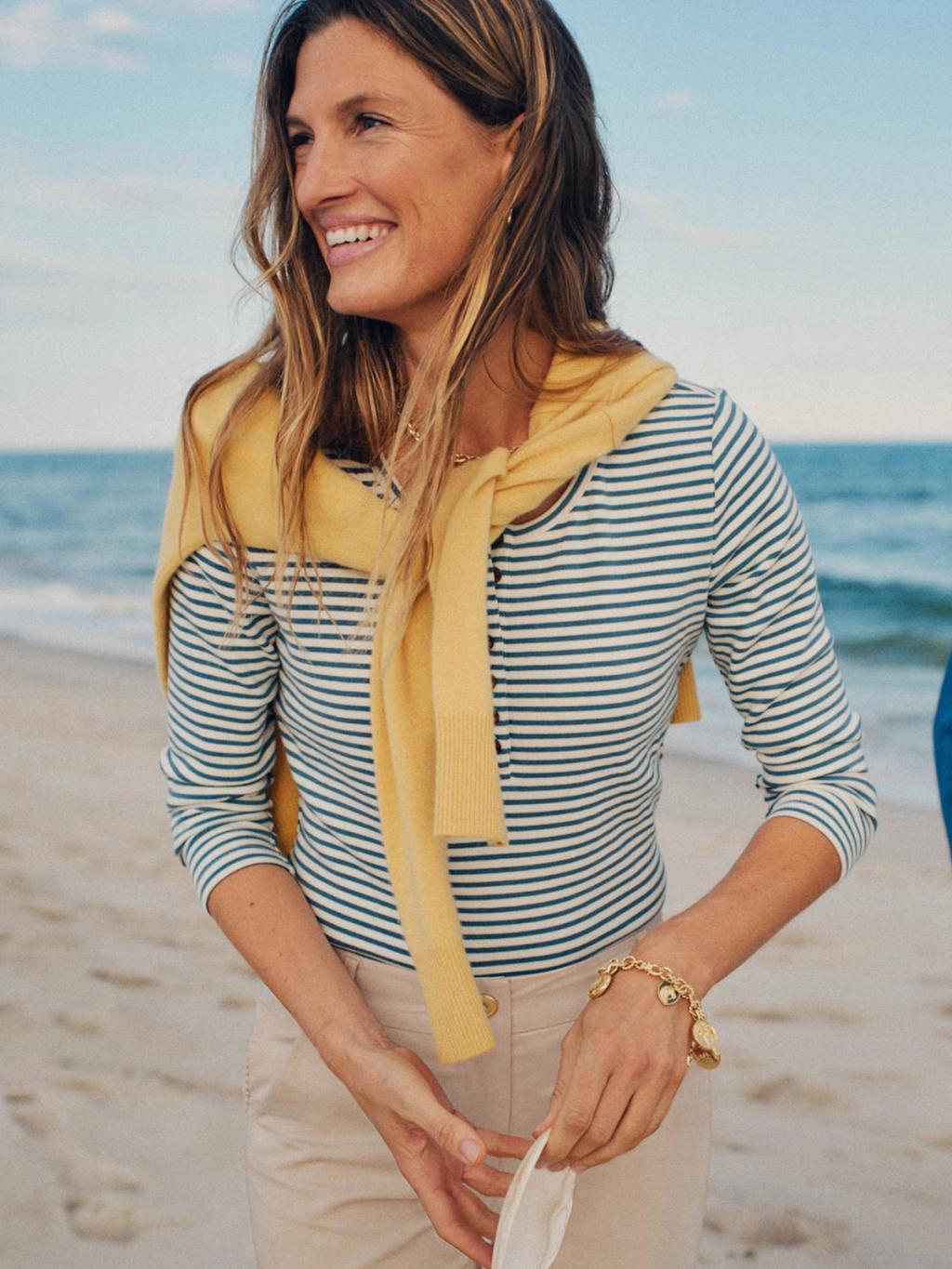 A smiling woman in a striped shirt and yellow sweater on a sandy beach.