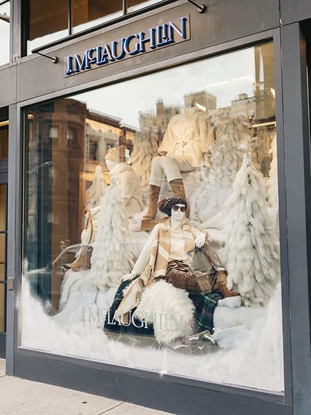Retail window display featuring winter-themed mannequins arranged among snow-covered props and white faux trees. The mannequins are dressed in neutral-toned winter clothing, including coats, scarves, and boots. The storefront sign above the window reads ‘J.McLaughlin.’ Reflections of nearby buildings appear in the glass.