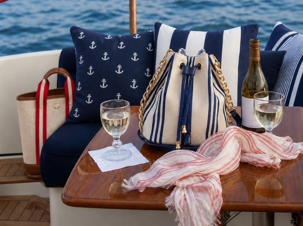 A nautical scene on a boat with wine, a striped bucket bag, and anchor-patterned cushions on a wooden table.