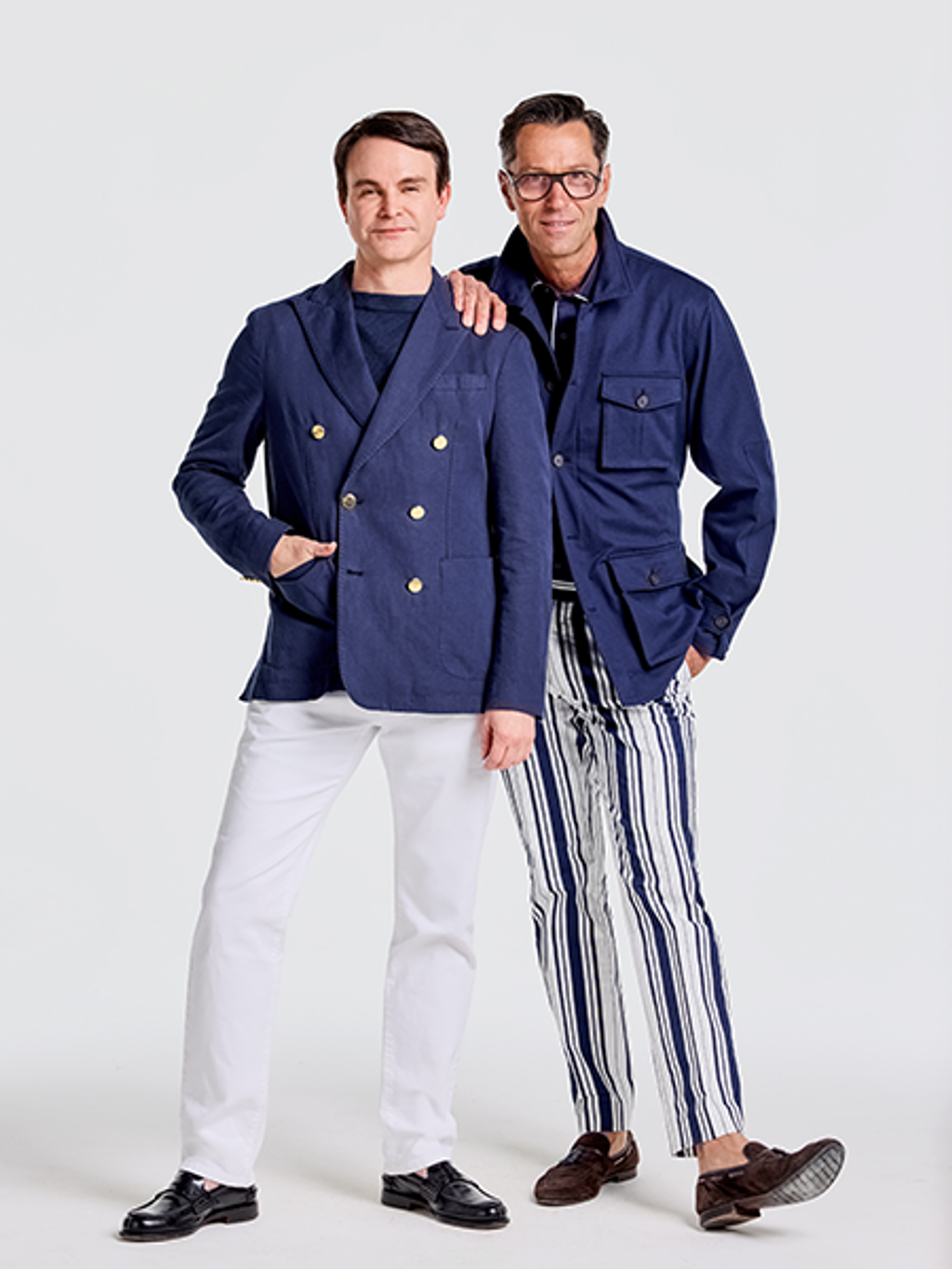 Two men; one in a navy double-breasted blazer and white pants, the other in glasses, a navy shirt-jacket, and striped pants, with his arm around the first man.