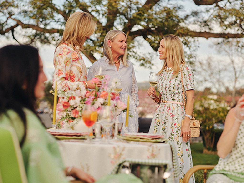 Three women in elegant dresses socialize around an outdoor table decorated with flowers and drinks.