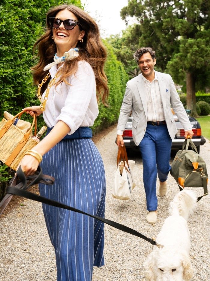 woman in navy pleated skirt and white linen shirt walking dogs down gravel driveway
