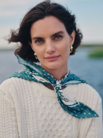 A woman with dark hair and blue eyes wears a cream cable-knit sweater and a teal patterned scarf.