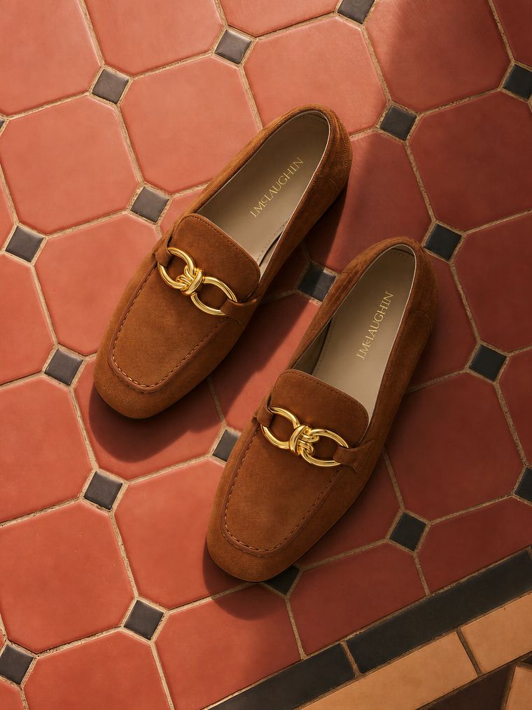 A pair of brown suede loafers with gold chain link hardware on a terracotta tile floor.