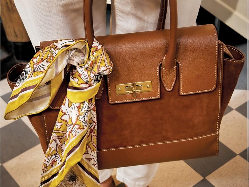 A person holds a brown leather handbag with a yellow floral scarf, wearing white pants and brown suede loafers on a checkered floor.
