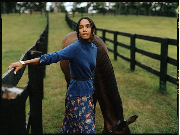 Female model in blue sweater, patterned skirt, and belt, posing with a brown horse.