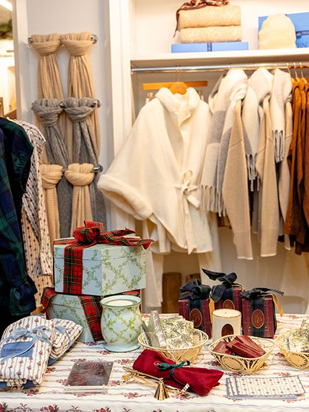 Interior view of a retail clothing store displaying merchandise on racks and shelves, with wrapped gift boxes and various items arranged on a table in the foreground.