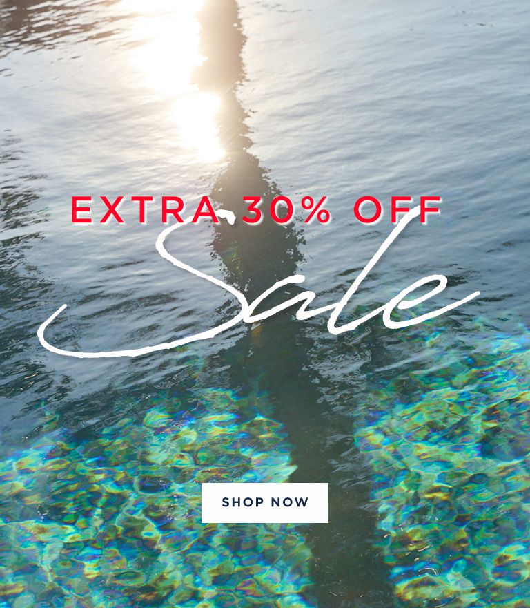 Split promotional image showing a woman in a white dress with black stripes carrying a cream and black striped sweater, standing outdoors with soft lighting on the left side. The right side features text over a water background with iridescent reflections reading 'EXTRA 30% OFF Sale' with a white 'SHOP NOW' button below.