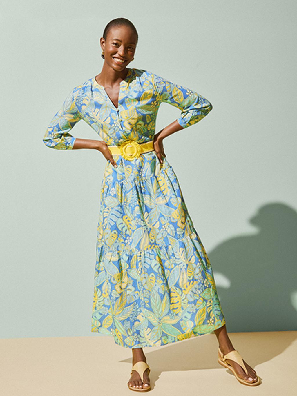Model in an ankle length blue/yellow floral dress with a matching yellow belt and tan sandals.