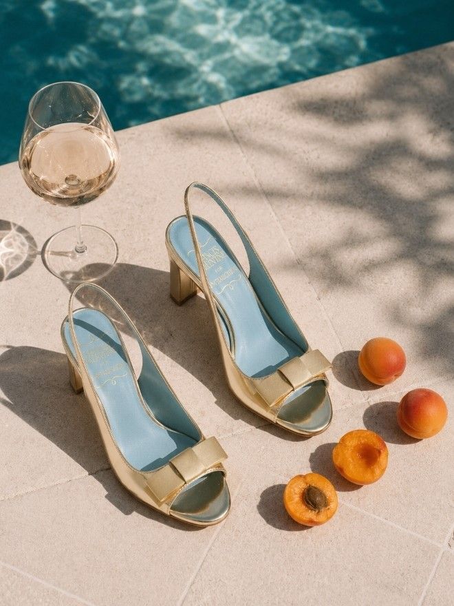 Golden slingback heels with blue insoles, a glass of white wine, and apricots on a poolside patio in dappled sunlight.