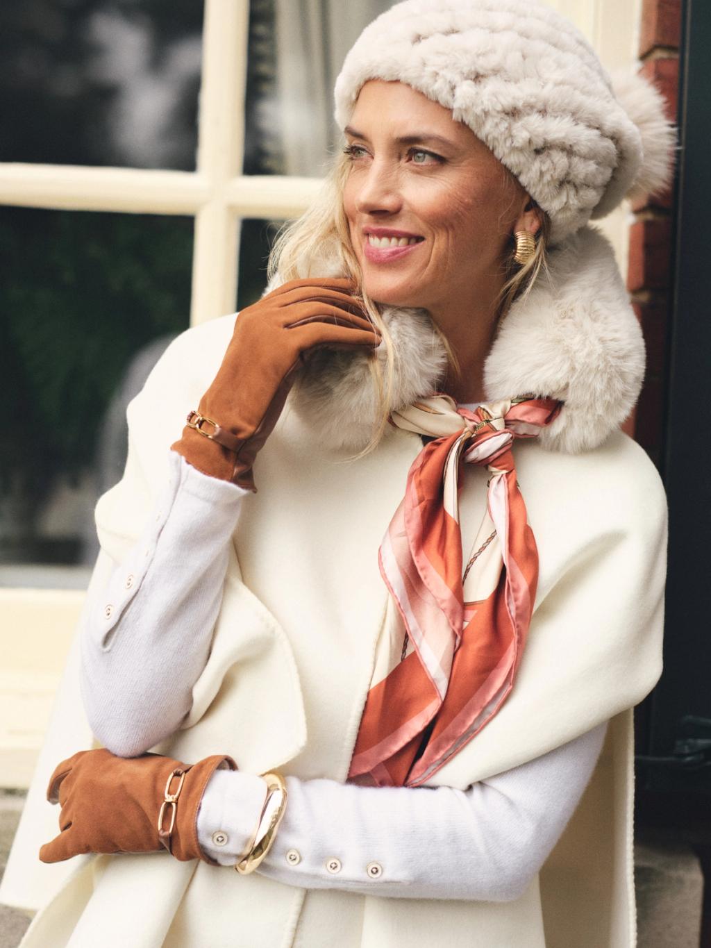 Smiling blonde woman in a white faux fur hat, white faux fur collar, cream cape, brown leather gloves, and a rust-patterned silk scarf.