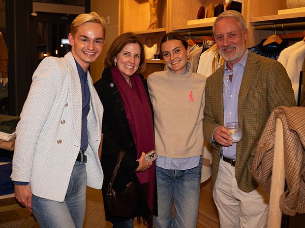Four people posing together in a clothing store, smiling at the camera. From left to right: a young man in a white textured blazer over a navy shirt, a woman in a black outfit with a burgundy scarf, a young woman in a beige sweatshirt with a pink ribbon logo and jeans, and an older man in a brown plaid blazer with a light blue shirt. Clothing racks and shelves are visible in the background.