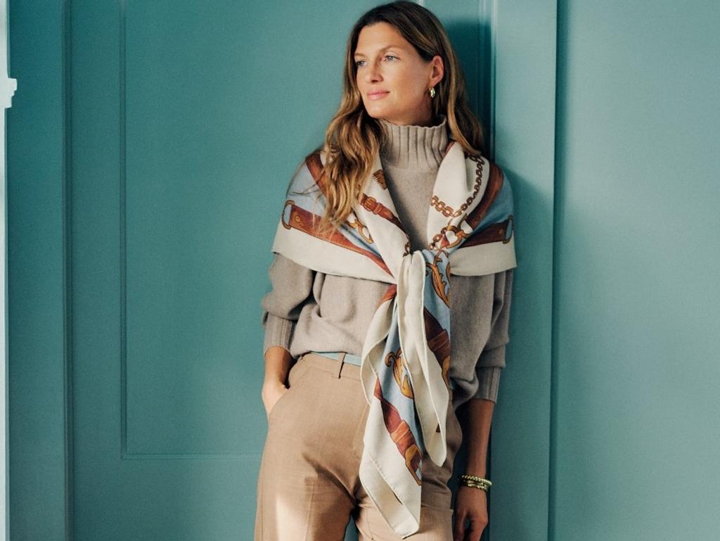A woman in a tan turtleneck, khaki pants, and a patterned scarf leans against a teal wall.