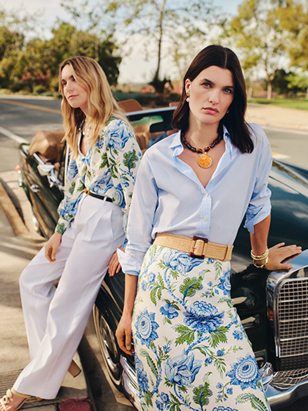 Two women in complementary blue and white outfits, featuring floral patterns, pose by a black vintage convertible car.