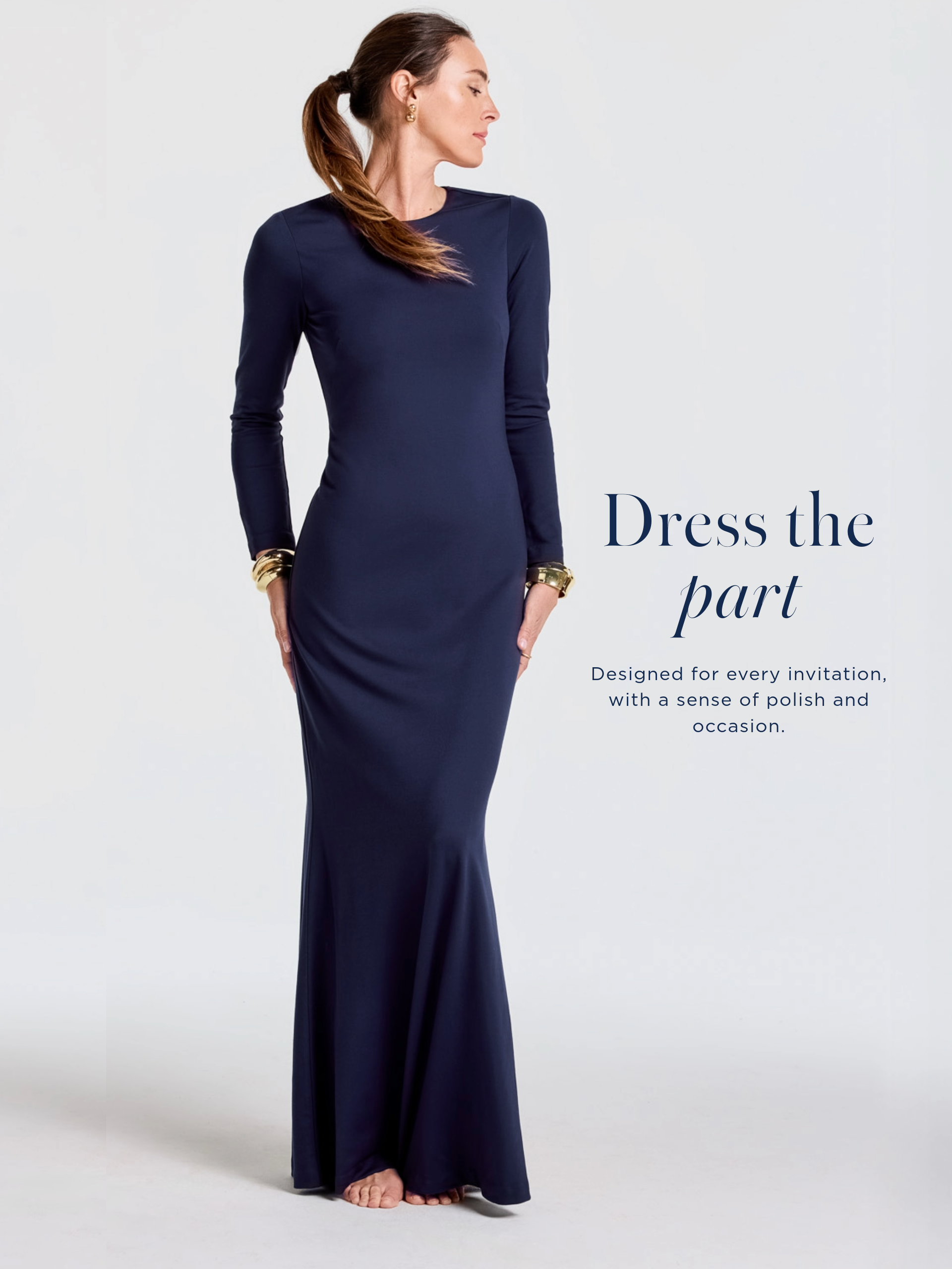 A woman models a form-fitting, long-sleeved navy blue gown with a flared hem, accessorized with gold earrings and bangles.