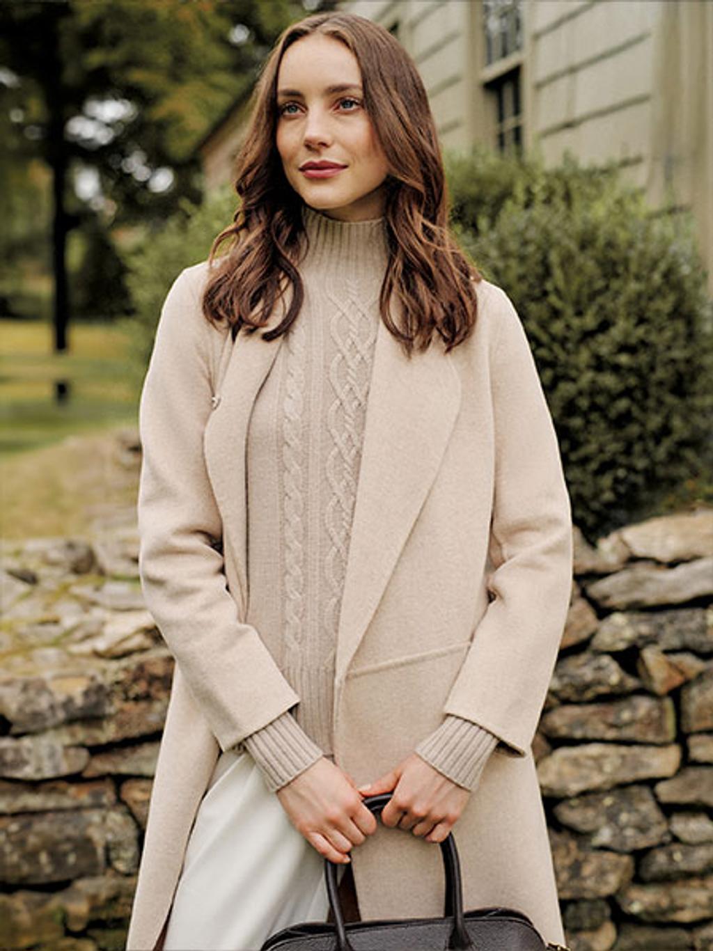 Model with brown hair wearing a cream wool coat and sweater holding a black handbag.