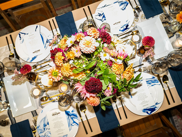 Overhead view of an elegantly set dining table featuring a lush floral centerpiece with dahlias in vibrant shades of burgundy, pink, yellow, orange, and white surrounded by green foliage. The table is set with white plates decorated with blue floral motifs, navy blue napkins, gold flatware, and wine glasses. Votive candles in gold holders are placed around the centerpiece. Menu cards are positioned at each place setting on the tan or beige table runner.