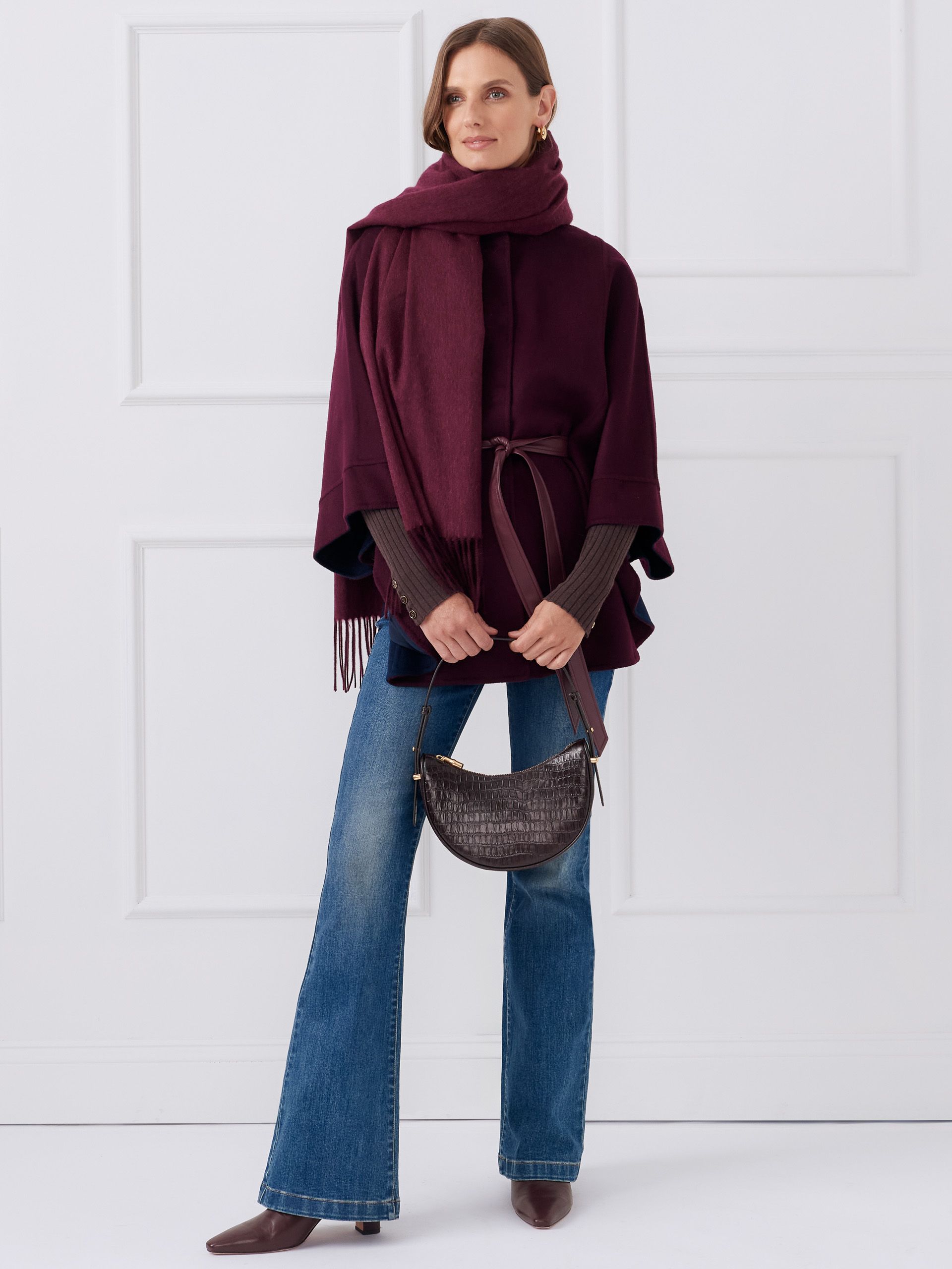 woman standing against white studio background wearing bootcut denim jeans a burgundy cape a burgundy cashmere sweater and a dark brown leather crescent shape bag
