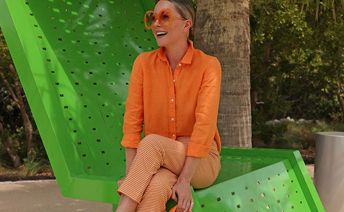 Influencer, Blaire Edie sitting on green chair wearing a bright orange Britt linen shirt, orange and white gingham pants.