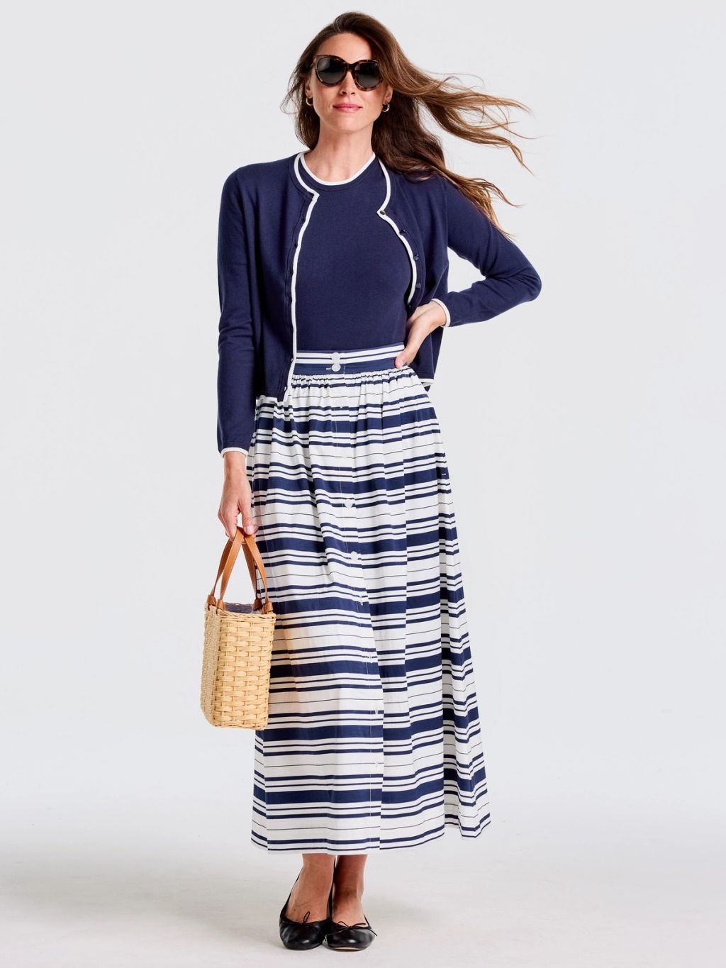 A woman in a navy top, white-trimmed navy cardigan, navy and white striped maxi skirt, sunglasses, and holding a straw bag.