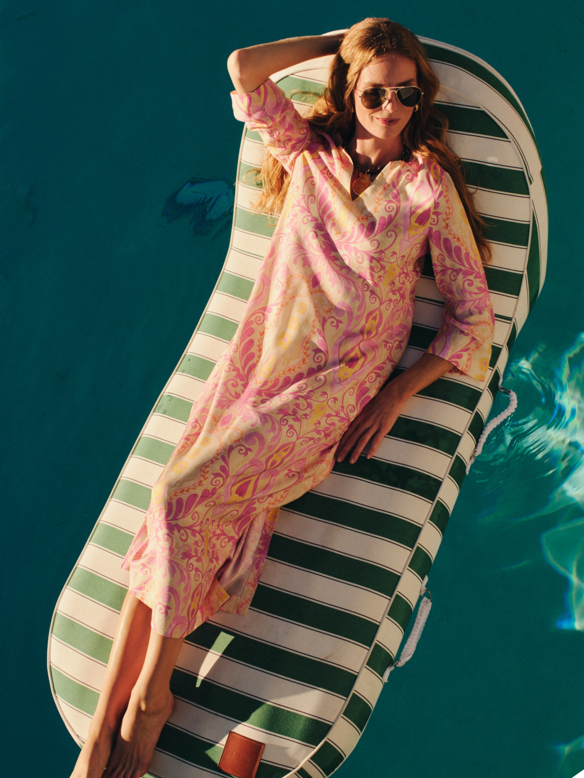 A woman in a pink patterned caftan and sunglasses reclines on a striped pool float in a blue pool.