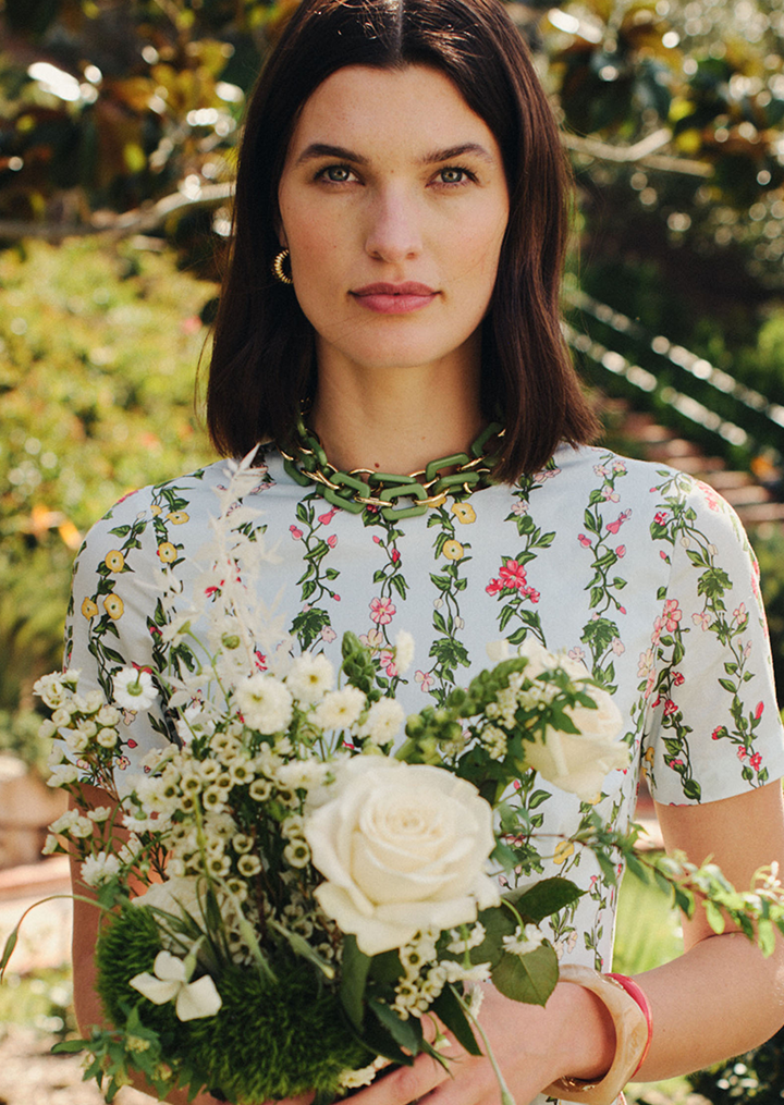 A woman in a light blue floral dress and green chain necklace holds a bouquet of white flowers with a white rose.