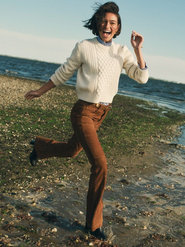 A joyful woman in a white cable-knit sweater and brown pants runs, splashing water on a muddy beach.