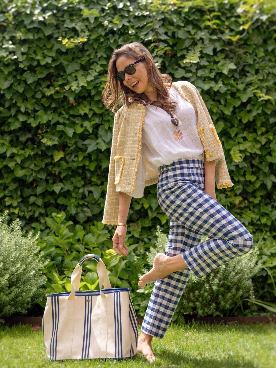 A smiling woman in sunglasses, a yellow tweed jacket, a white top, and blue gingham pants poses barefoot on grass next to a striped tote bag.