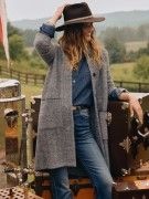 woman standing outside leaning on a steamer trunk wearing jeans, a denim shirt, a cowboy hat and a gray long cardigan coat