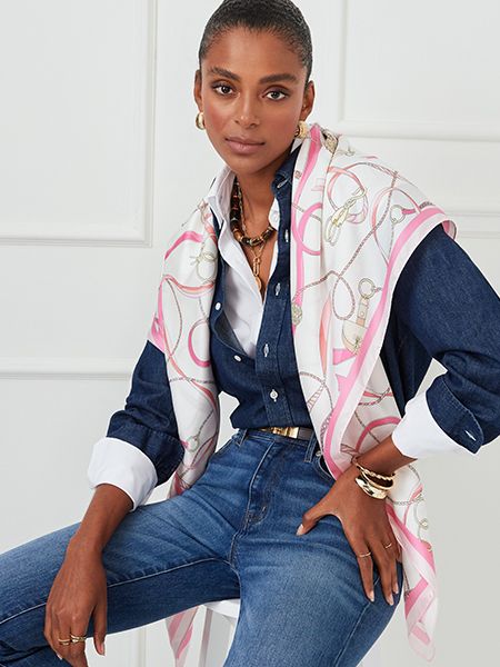 Woman wearing a pink and white chain-print scarf over a navy blue button-up shirt, white undershirt, and blue jeans, accessorized with gold jewelry including hoop earrings, a statement necklace, and multiple rings, seated against a white paneled wall