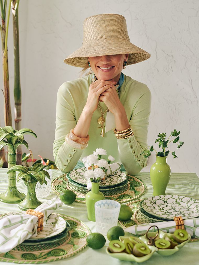 woman seated at dining table with green decor wearing a green sweater and a large sun hat. 
